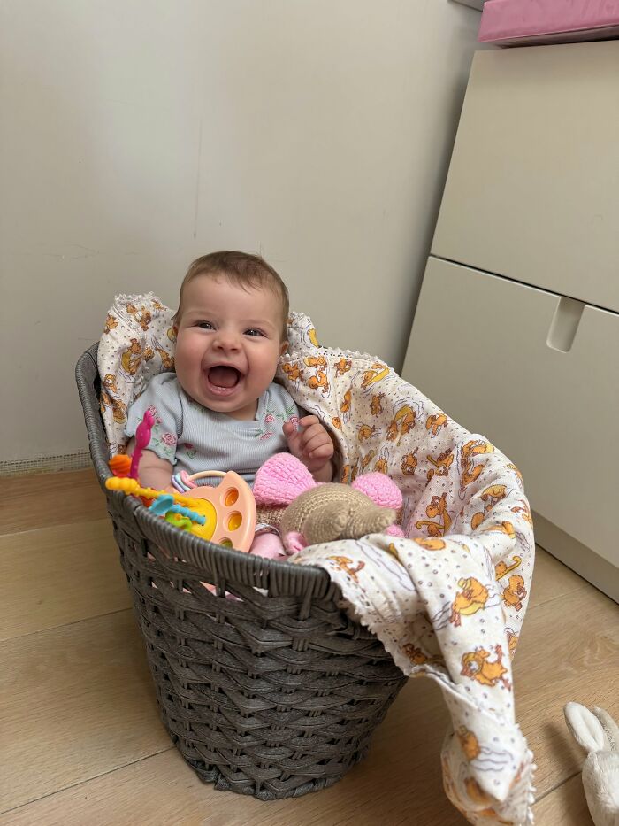 A joyful baby sitting in a grey woven basket, surrounded by a patterned blanket and toys, representing the rewarding side of motherhood.