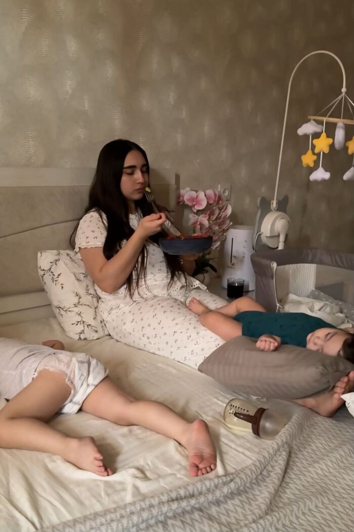 A mom eating while her two children sleep on the bed beside her, capturing the reality that motherhood is not easy.