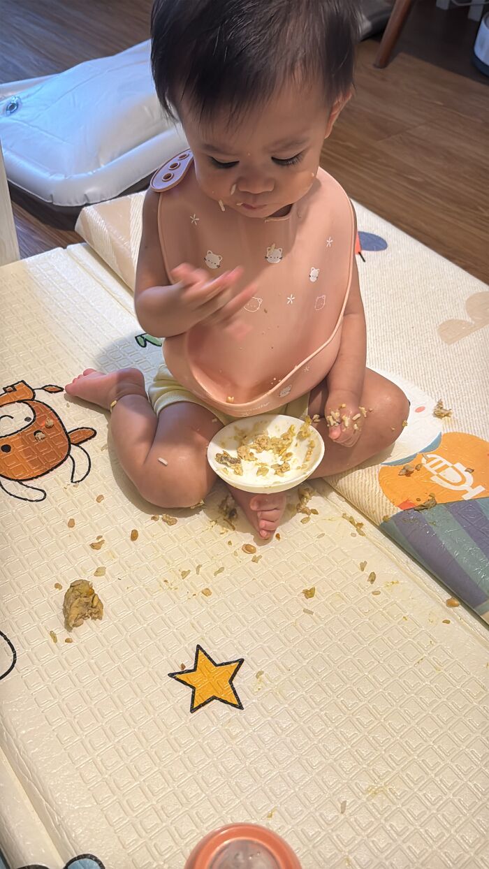A baby in a pink bib sits on a mat, making a mess while eating, highlighting the rewarding yet not easy journey of motherhood.
