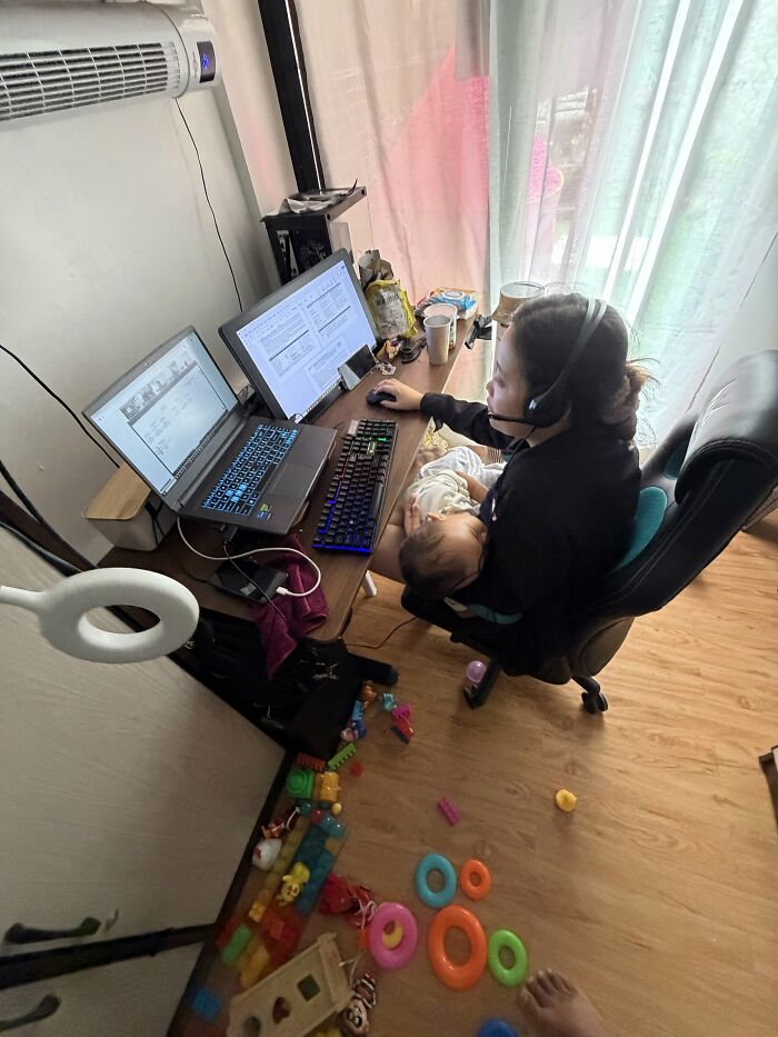 A mom works at a computer desk while holding her baby, highlighting the challenging but fulfilling reality of motherhood.
