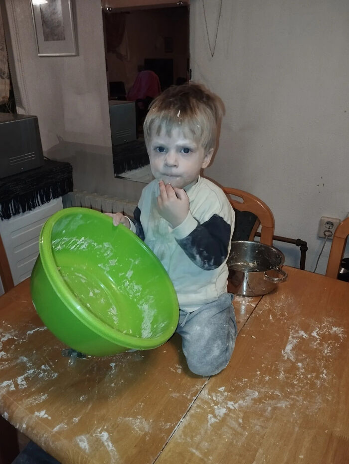 A young child sits on a table covered in flour next to a green bowl, embodying the challenging yet fulfilling journey of motherhood.