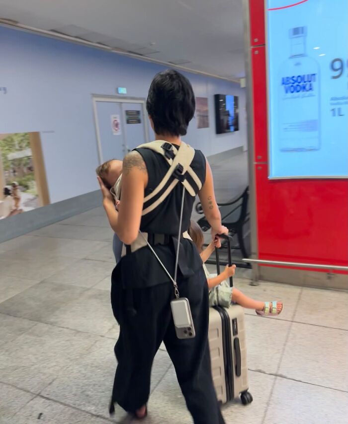 A mom with a baby in a carrier and a child riding on luggage at an airport, depicting the rewarding but challenging journey of motherhood.