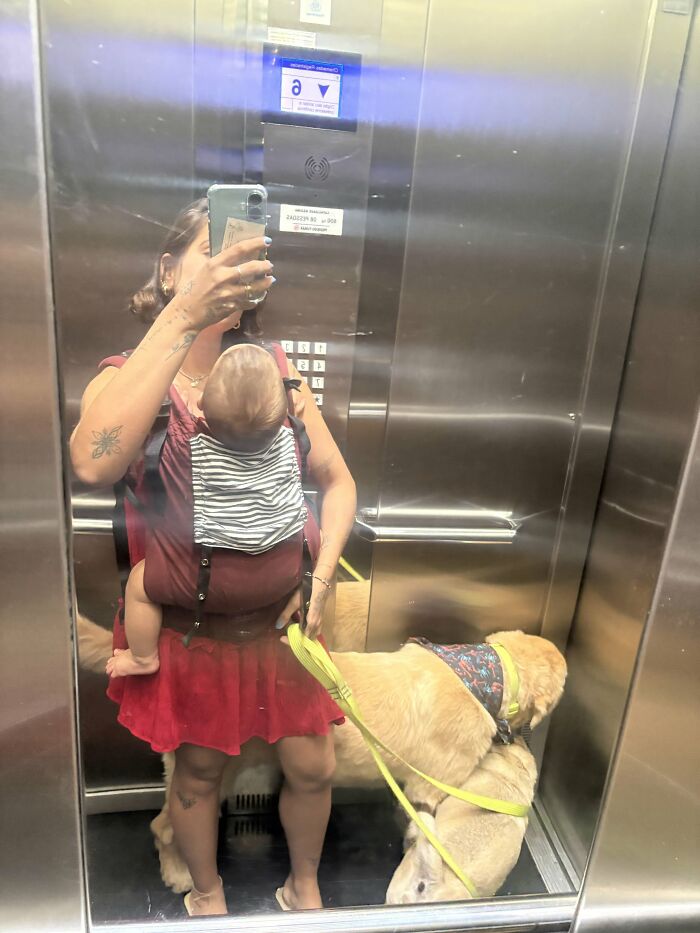 A mom in an elevator with a baby in a carrier and a dog on a leash, illustrating the rewarding and joyful yet not easy life of motherhood.