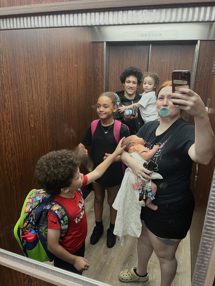 A mom takes a family selfie in an elevator, showcasing the rewarding and joyful but not easy aspects of motherhood.