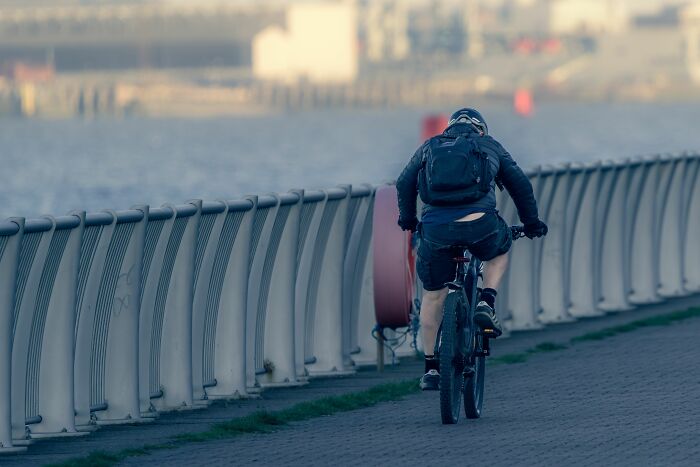 A cyclist riding along a paved path next to a body of water, an absurd yet common sight for daily commuters.