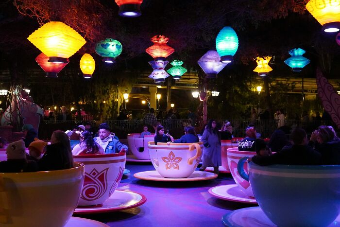 People riding in teacup rides under colorful lanterns, a hilarious and absurd scene at an amusement park.