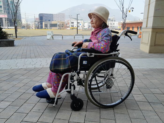An elderly woman in a wheelchair sits outdoors, witnessing firsthand a calm moment.