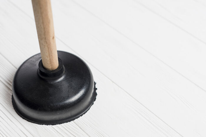 A black rubber plunger with a wooden handle on a white wooden floor, an absurd and hilarious firsthand object.