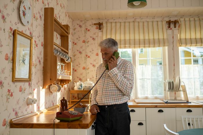 An elderly man talks on a retro phone in a floral kitchen. A firsthand witnessed scene of potentially absurd and hilarious old tech.