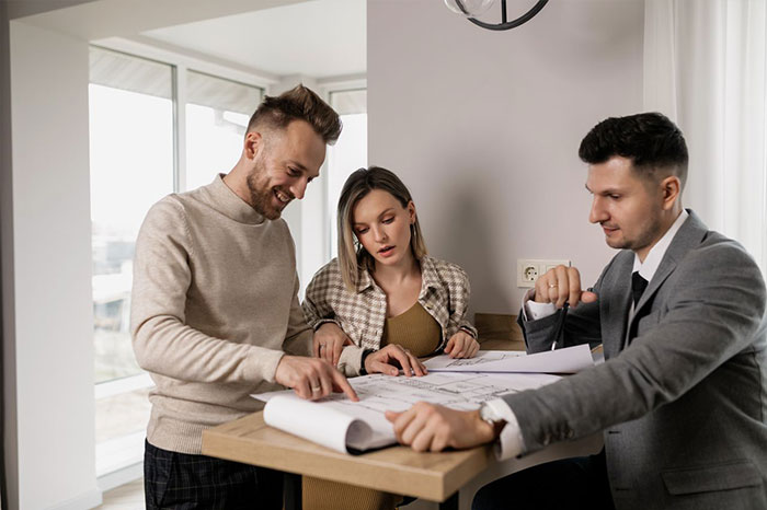 A couple reviews blueprints with a professional, perhaps discussing property or a secret scheme related to an HOA.