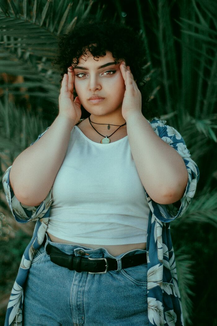 A young woman with curly hair looks at the camera, her hands on her temples. She is one of the interesting neighbors.