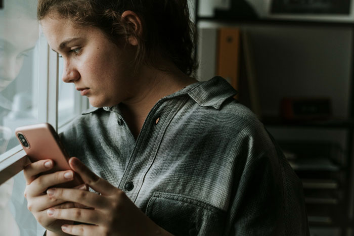 A young woman looks out a window, holding a phone, contemplating her violent fight with sister as a teen.