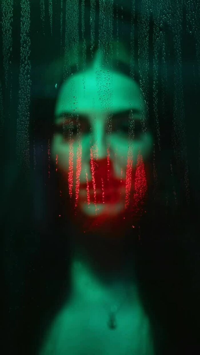A blurred woman's face through a rainy window, illuminated by green and red neon lights, creating a moody cinematic portrait.