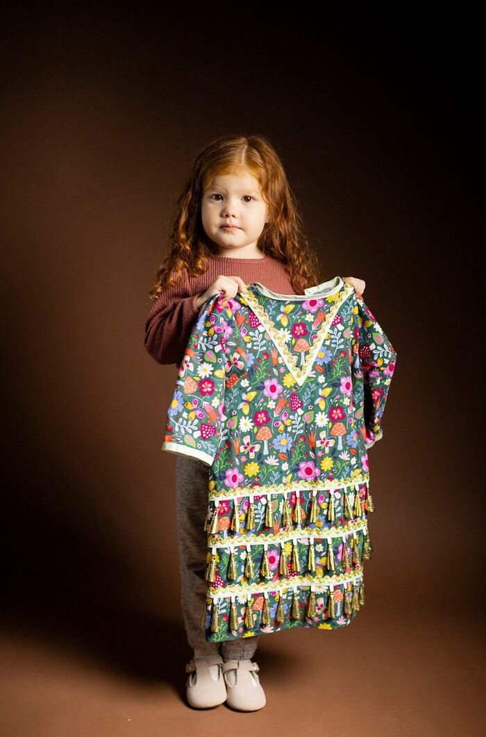 A small child with long, wavy natural redheads hair, holding up a colorful dress.