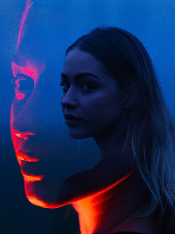 A double exposure cinematic portrait of a woman, with one face illuminated by red neon lights and the other in blue.