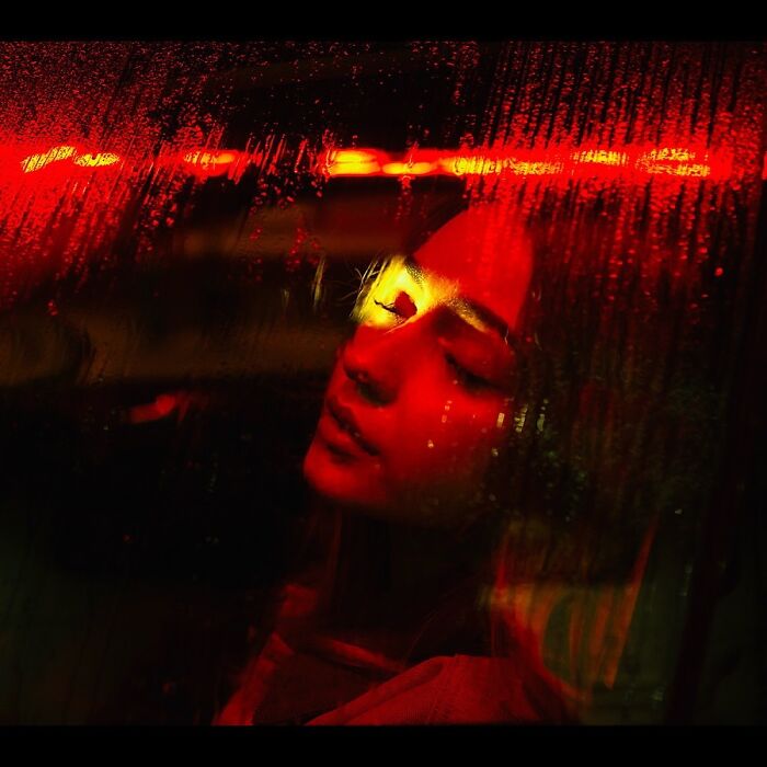 A woman with closed eyes, illuminated by red neon lights through a wet window, creating a cinematic portrait.