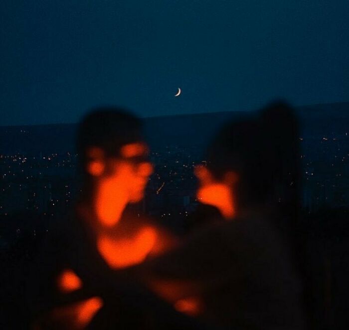 A couple in silhouette, bathed in red neon lights, against a dark sky with a crescent moon and city lights below. This is an example of cinematic portraits.