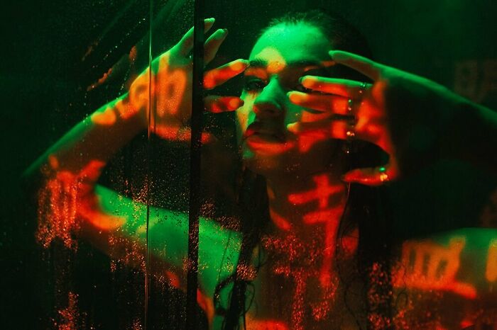 A man with long hair, illuminated by green and red neon lights, presses his hands against a wet glass, creating a cinematic portrait.