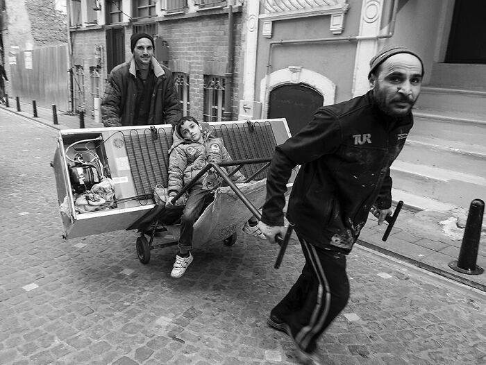 Candid moments from the streets, two men and a boy on a cart with a refrigerator, walking down a narrow street in black and white.