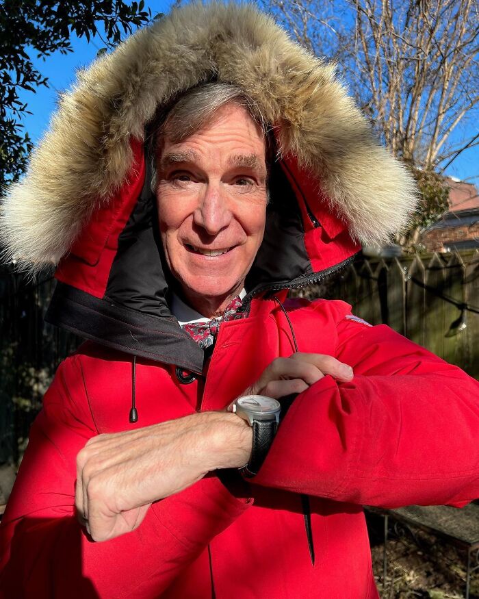 Bill Nye smiles in a red fur-hooded coat, checking his watch, perhaps revealing insights into dating a celebrity.