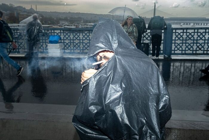 A candid moment from the streets: a person wearing a black plastic bag smokes in the rain.