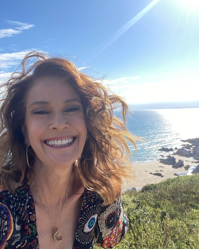 A smiling celebrity woman taking a selfie outdoors with a sunny beach and ocean in the background, showing what dating a celebrity is like.