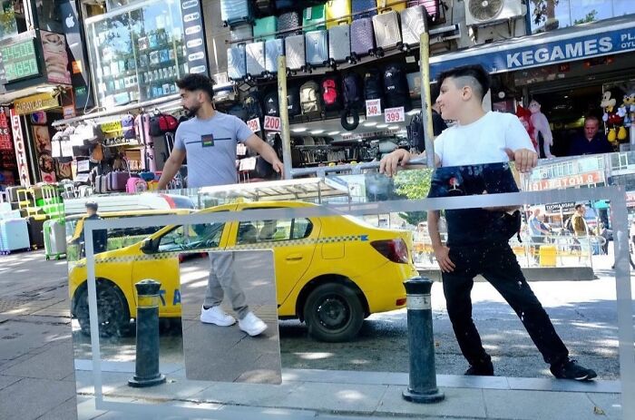 A candid moment from the streets: two young men pose with reflections of a yellow taxi in mirrors on a city street.