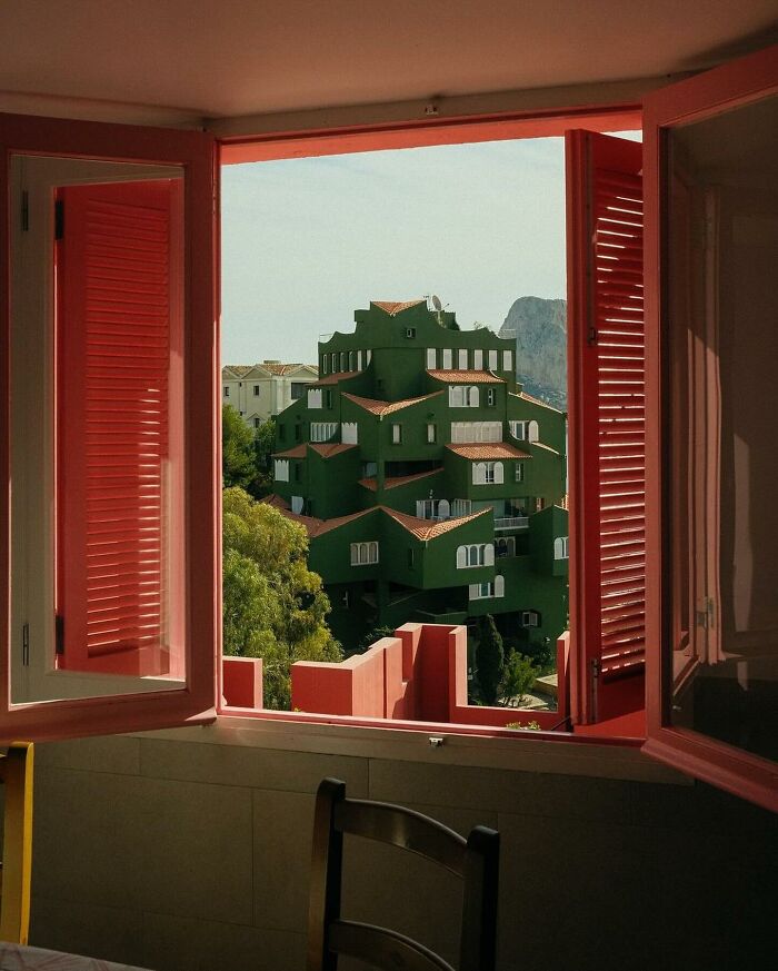 A brilliant and beautiful green terraced building with red roofs viewed through an open window with red shutters, a design highlight.