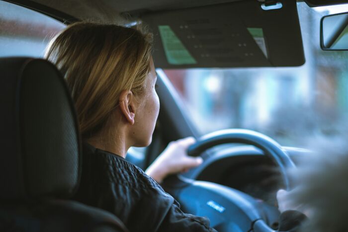 A blonde woman driving, looking out the windshield, possibly approaching a drive-through. Features a customer in a car.