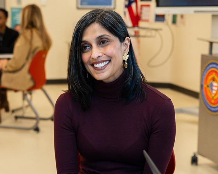 A woman with dark hair and a maroon turtleneck smiles, looking to the side in a room. A woman with dark hair and a maroon turtleneck smiles, looking to the side in a room.