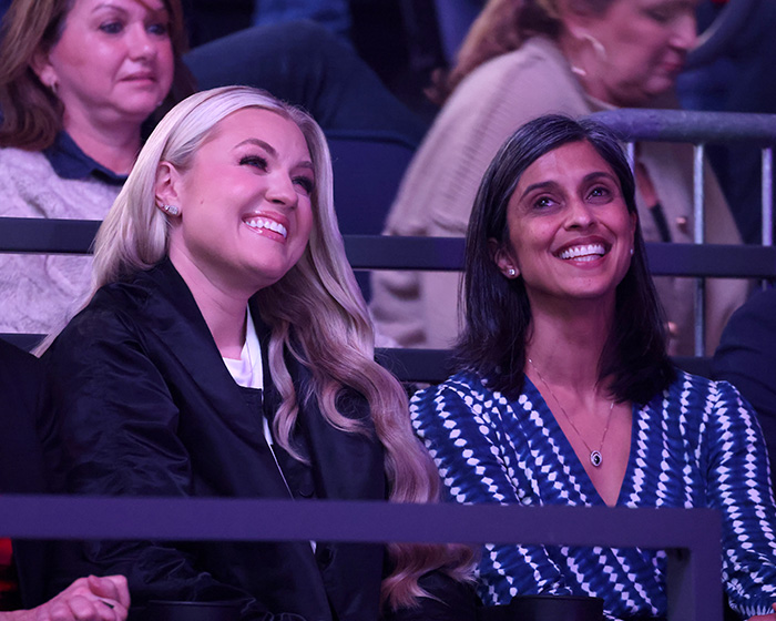 Two women, one with blonde hair, one with dark hair, smiling while sitting in an audience. Two women, one with blonde hair, one with dark hair, smiling while sitting in an audience.