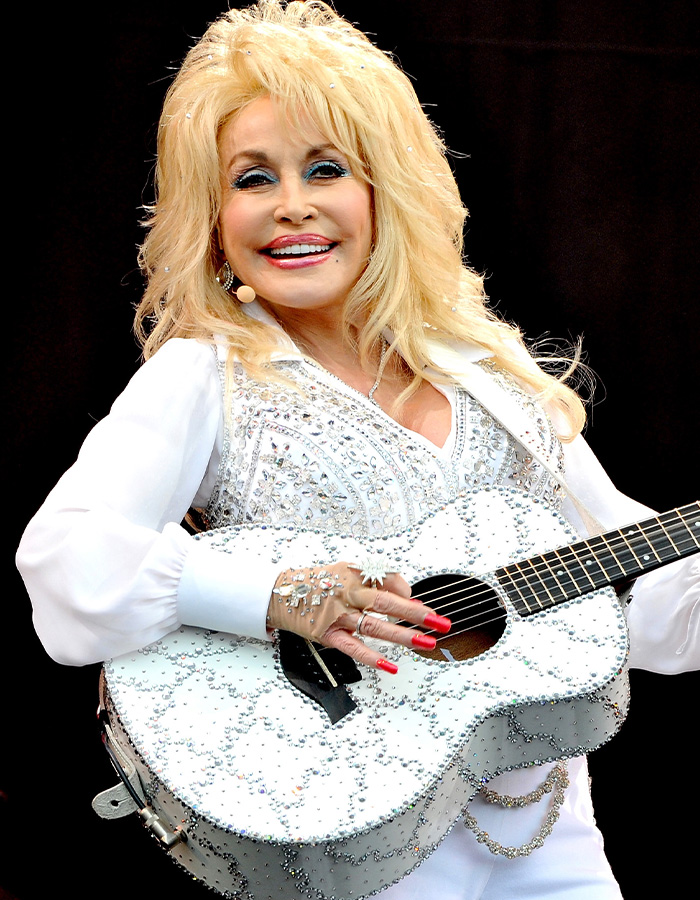 Dolly Parton on stage, holding a rhinestone-studded guitar, smiling at the audience. Dolly Parton on stage, holding a rhinestone-studded guitar, smiling at the audience.