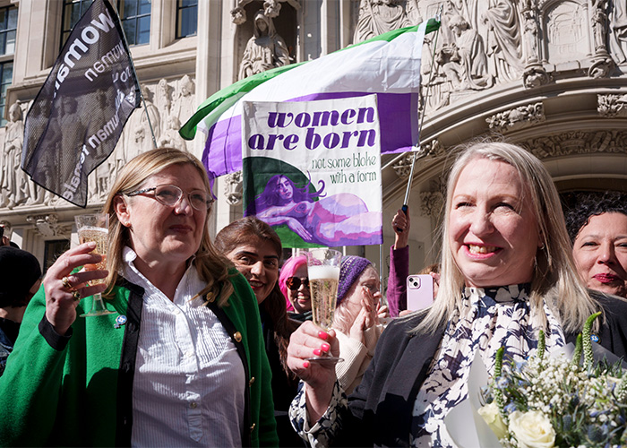 Two women smiling and holding champagne glasses, celebrating in front of a building with signs about women.