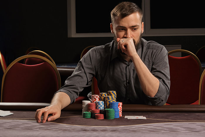A man with a beard sits at a poker table, contemplating his stack of chips, possibly doing a weird job.