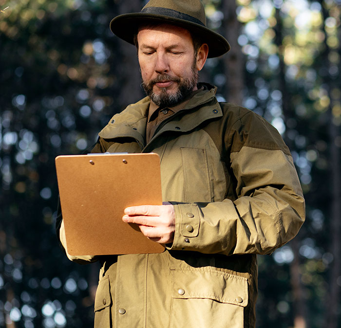 A man in a hat and jacket, holding a clipboard in a forest, for weird jobs.