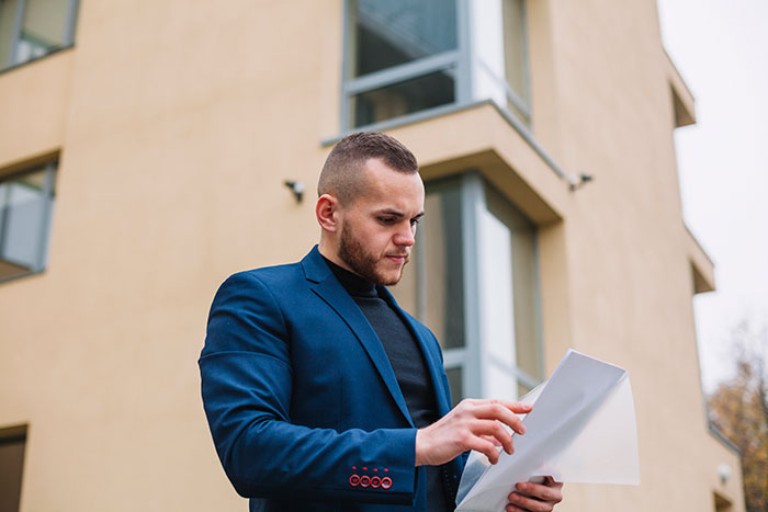 A man in a blue suit looks at papers outside a modern building, possibly related to some weird jobs.