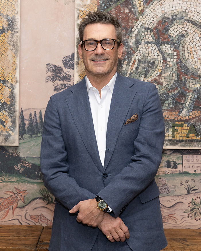 Jon Hamm, expecting first child, in a blue suit and glasses, smiling against a mosaic background. Jon Hamm, expecting first child, in a blue suit and glasses, smiling against a mosaic background.