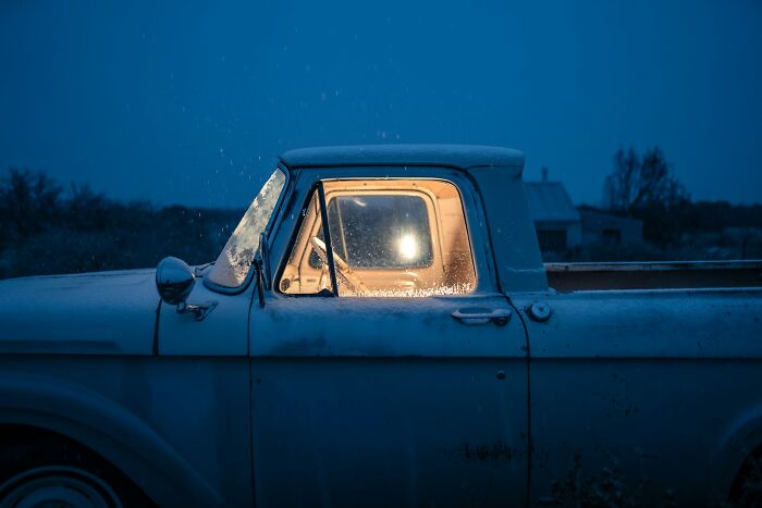 An old truck at dusk with a light on inside, covered in a light dusting of snow, for scary accurate true stories.