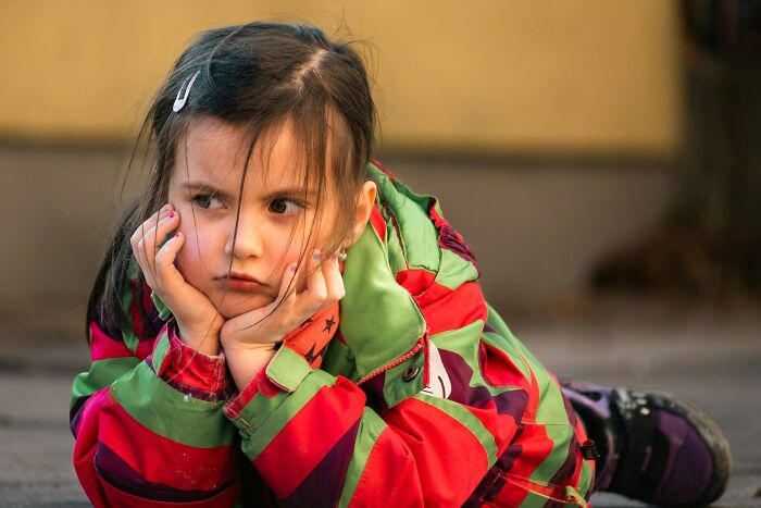 A sad young girl in a colorful jacket sits on the ground, reflecting on a horrible parent.