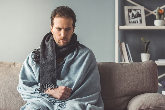 A man wrapped in a blanket and scarf sitting on a couch, experiencing cold due to a landlord refusing to fix heating. A man wrapped in a blanket and scarf sitting on a couch, experiencing cold due to a landlord refusing to fix heating.