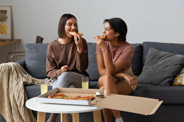 Two young women sharing pizza on a couch, representing the personal lounge aspect of the story about a sis. Two young women sharing pizza on a couch, representing the personal lounge aspect of the story about a sis.