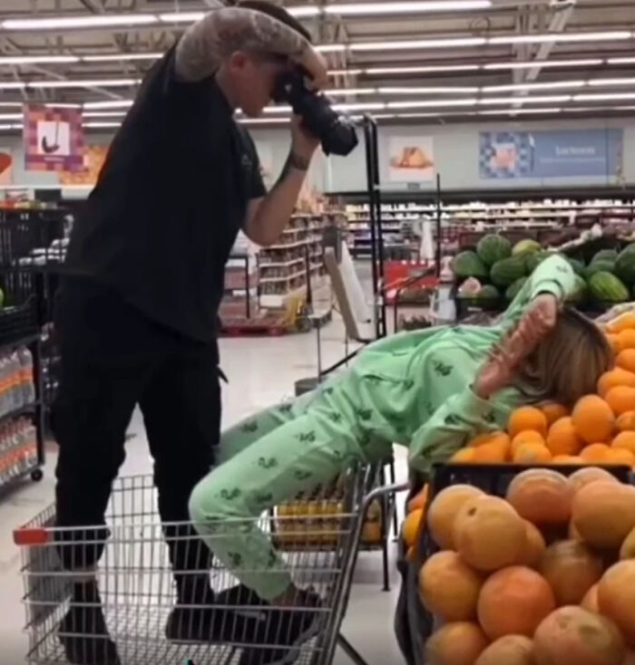 An entitled photographer takes pictures of a woman posing in a supermarket cart, surrounded by produce.