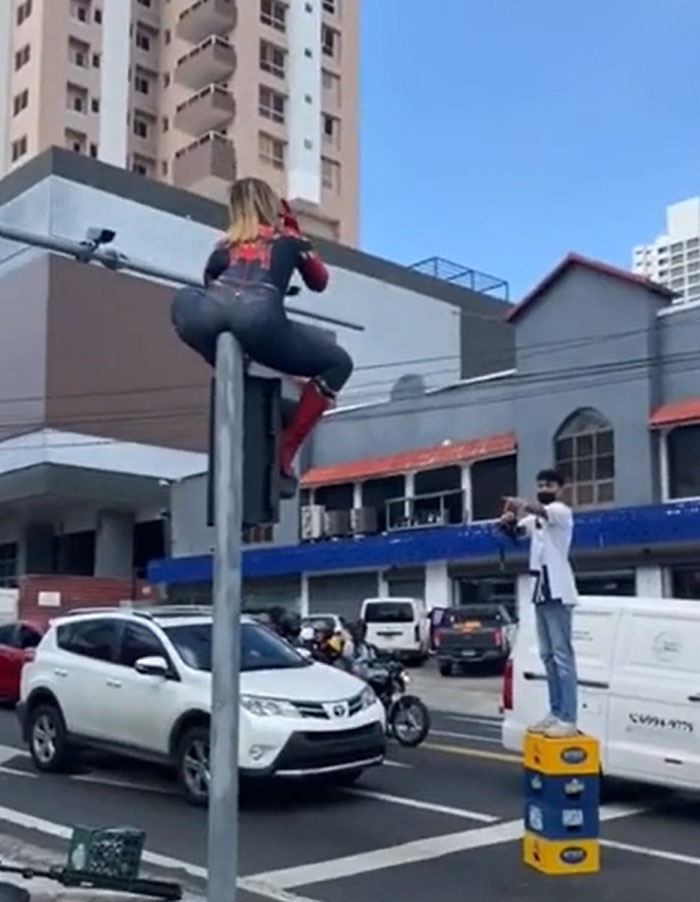 Entitled woman in a Spider-Man suit sitting on a traffic light pole, posing for photos in public.