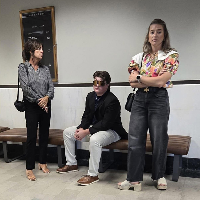 An Oklahoma teen in sunglasses seated on a bench with two women in a hallway, awaiting legal proceedings for brutal crimes.