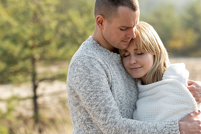 A husband embraces his wife, who is wrapped in a blanket, both closing their eyes in a moment of peace. A husband embraces his wife, who is wrapped in a blanket, both closing their eyes in a moment of peace.