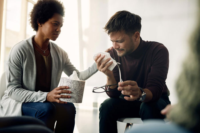 Man mourns his marriage, crying into a tissue while a woman offers comfort during a therapy session.