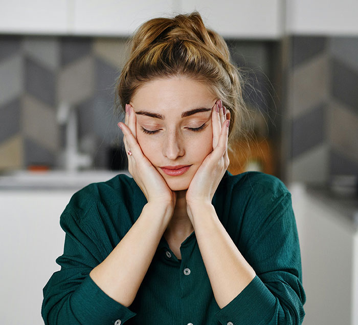 A young woman in a green shirt, eyes closed, holding her head in her hands, depicting the emotional impact of mental load. A young woman in a green shirt, eyes closed, holding her head in her hands, depicting the emotional impact of mental load.