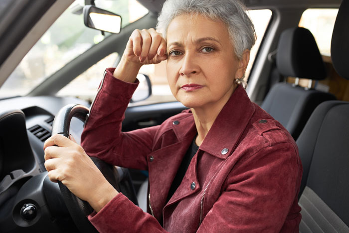 A senior woman with gray hair sits in a car, looking sternly at the camera, conveying a selfish mom's concern about her son's debt. A senior woman with gray hair sits in a car, looking sternly at the camera, conveying a selfish mom's concern about her son's debt.