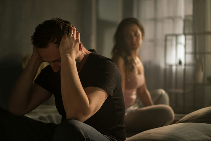 A distraught cheating husband sits on the bed with his wife in the background, showing marital strain. A distraught cheating husband sits on the bed with his wife in the background, showing marital strain.