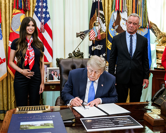 Mystery woman behind Trump with a strange gesture as he signs documents at a desk.